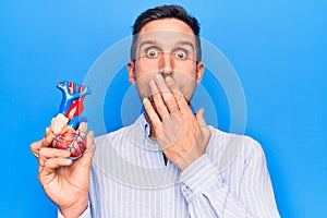 Young handsome man holding heart organ with veins and arteries over blule background covering mouth with hand, shocked and afraid