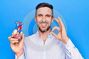 Young handsome man holding heart organ with veins and arteries over blule background doing ok sign with fingers, smiling friendly