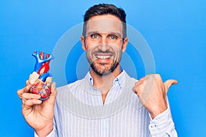 Young handsome man holding heart organ with veins and arteries over blule background pointing thumb up to the side smiling happy