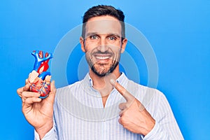 Young handsome man holding heart organ with veins and arteries over blule background smiling happy pointing with hand and finger