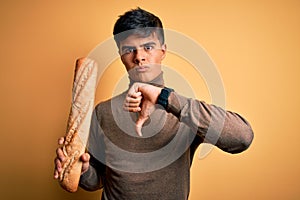 Young handsome man holding homemade fresh bread over isolated yellow background with angry face, negative sign showing dislike