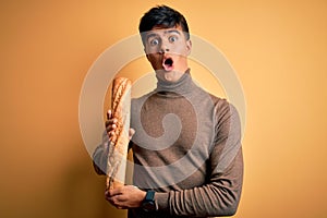 Young handsome man holding homemade fresh bread over isolated yellow background scared in shock with a surprise face, afraid and