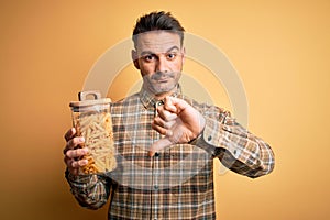 Young handsome man holding jar with dry italian pasta macaroni over yellow background with angry face, negative sign showing