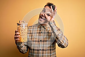 Young handsome man holding jar with dry italian pasta macaroni over yellow background with happy face smiling doing ok sign with