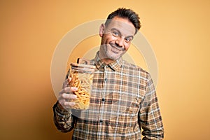 Young handsome man holding jar with dry italian pasta macaroni over yellow background with a happy face standing and smiling with