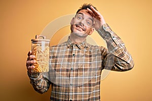 Young handsome man holding jar with dry italian pasta macaroni over yellow background stressed with hand on head, shocked with