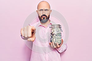 Young handsome man holding jar with savings pointing with finger to the camera and to you, confident gesture looking serious