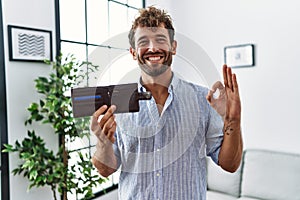 Young handsome man holding leather wallet doing ok sign with fingers, smiling friendly gesturing excellent symbol