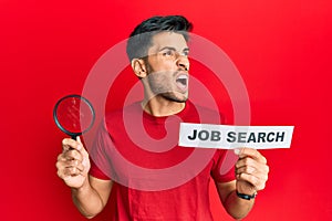 Young handsome man holding magnifying glass for job search angry and mad screaming frustrated and furious, shouting with anger