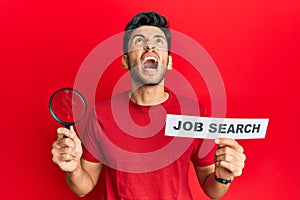 Young handsome man holding magnifying glass for job search angry and mad screaming frustrated and furious, shouting with anger