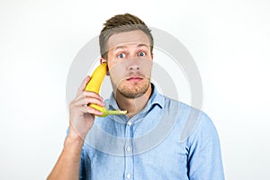 Young handsome man holding one fresh banana as a phone near his ear looks suspicious on isolated white background