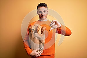 Young handsome man holding paper bag with bread over isolated yellow background with angry face, negative sign showing dislike