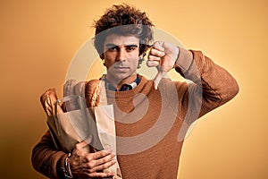 Young handsome man holding paper bag with bread standing over isolated yellow background with angry face, negative sign showing