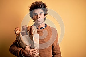 Young handsome man holding paper bag with bread standing over isolated yellow background with a confident expression on smart face