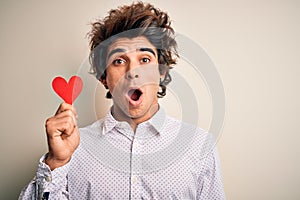 Young handsome man holding paper heart standing over isolated white background scared in shock with a surprise face, afraid and