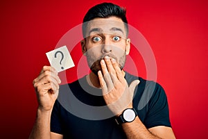 Young handsome man holding paper with question mark symbol over red background cover mouth with hand shocked with shame for