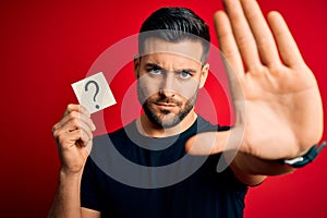 Young handsome man holding paper with question mark symbol over red background with open hand doing stop sign with serious and