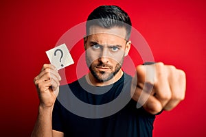 Young handsome man holding paper with question mark symbol over red background pointing with finger to the camera and to you, hand