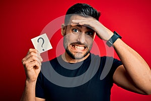 Young handsome man holding paper with question mark symbol over red background stressed with hand on head, shocked with shame and