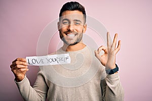 Young handsome man holding paper with self love message over pink background doing ok sign with fingers, excellent symbol