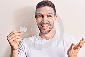 Young handsome man holding piece of puzzle standing over isolated white background celebrating achievement with happy smile and