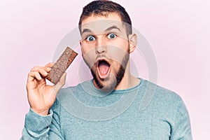 Young handsome man holding protein bar scared and amazed with open mouth for surprise, disbelief face