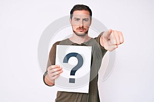 Young handsome man holding question mark pointing with finger to the camera and to you, confident gesture looking serious