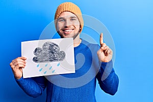 Young handsome man holding rain draw smiling with an idea or question pointing finger with happy face, number one