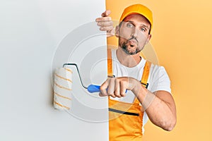 Young handsome man holding roller painter over white banner looking at the camera blowing a kiss being lovely and sexy