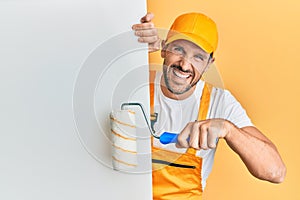 Young handsome man holding roller painter over white banner smiling and laughing hard out loud because funny crazy joke