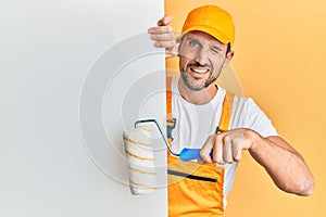 Young handsome man holding roller painter over white banner winking looking at the camera with sexy expression, cheerful and happy