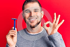 Young handsome man holding shave razor doing ok sign with fingers, smiling friendly gesturing excellent symbol