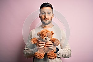Young handsome man holding teddy bear standing over isolated pink background with a confident expression on smart face thinking