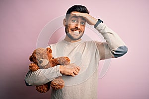 Young handsome man holding teddy bear standing over isolated pink background stressed with hand on head, shocked with shame and