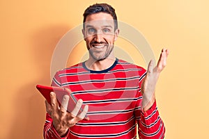 Young handsome man holding touchpad standing over isolated yellow background celebrating achievement with happy smile and winner