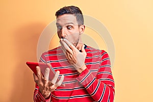 Young handsome man holding touchpad standing over isolated yellow background covering mouth with hand, shocked and afraid for