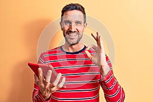 Young handsome man holding touchpad standing over isolated yellow background doing ok sign with fingers, smiling friendly