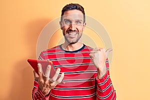 Young handsome man holding touchpad standing over isolated yellow background pointing thumb up to the side smiling happy with open