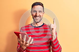 Young handsome man holding touchpad standing over isolated yellow background smiling happy pointing with hand and finger to the