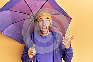 Young handsome man holding umbrella pointing thumb up to the side smiling happy with open mouth