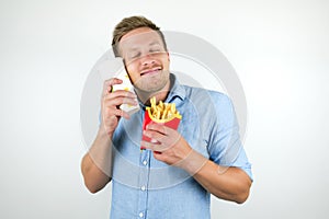 Young handsome man holds paper cup with soda near his satisfied face while eating eats french fries from fast food