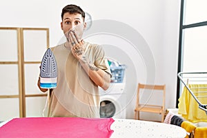 Young handsome man ironing clothes at home covering mouth with hand, shocked and afraid for mistake