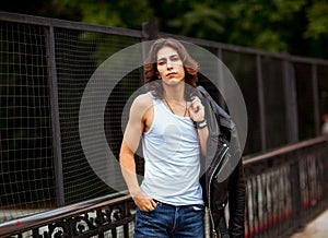 A young, handsome man with long, dark hair, in a white T-shirt and jeans, walks on the bridge