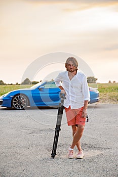 A young handsome man with long hair stands with a tripod somewhere on a country road, the photographer artist arrived in