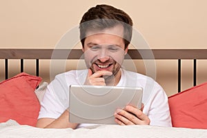 Young handsome man lying in bed and using tablet computer or reading an ebook