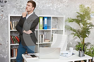 Young handsome man manager standing near his working place in office looking thoughtful at the end of day. multitasking, work