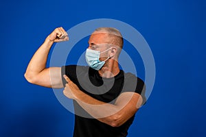 Young handsome man in medical mask and black t-shirt isolated on blue background. Strong person showing arm muscle