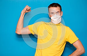 Young handsome man in medical mask and yellow t-shirt isolated on blue background. Strong person showing arm muscle, confident and