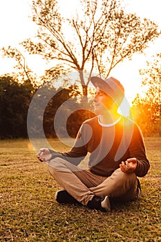 Young handsome man in a meditative position sits on a lawn on a background of forest and sunset. Concept of freedom relaxation.