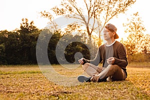 Young handsome man in a meditative position sits on a lawn on a background of forest and sunset. Concept of freedom relaxation.